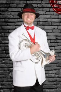 Image of a man smiling holding a trumpet. Wearing a white blazer and black fedora.